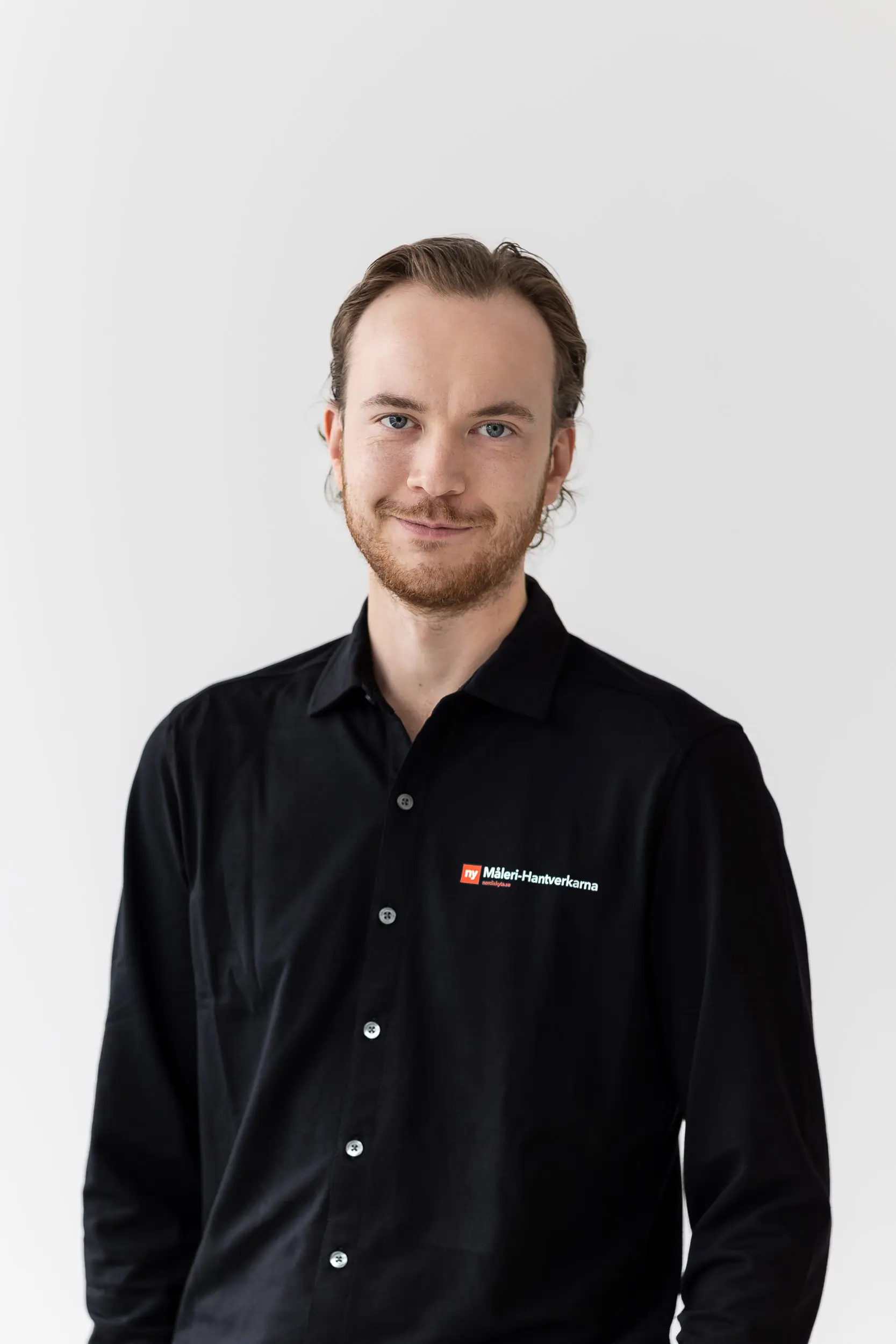 En man med ljusbrunt hår och vältrimmat skägg ler lätt. Han är klädd i en svart skjorta med Mälar-Hantverkarn-logotyp och står mot en vit bakgrund.