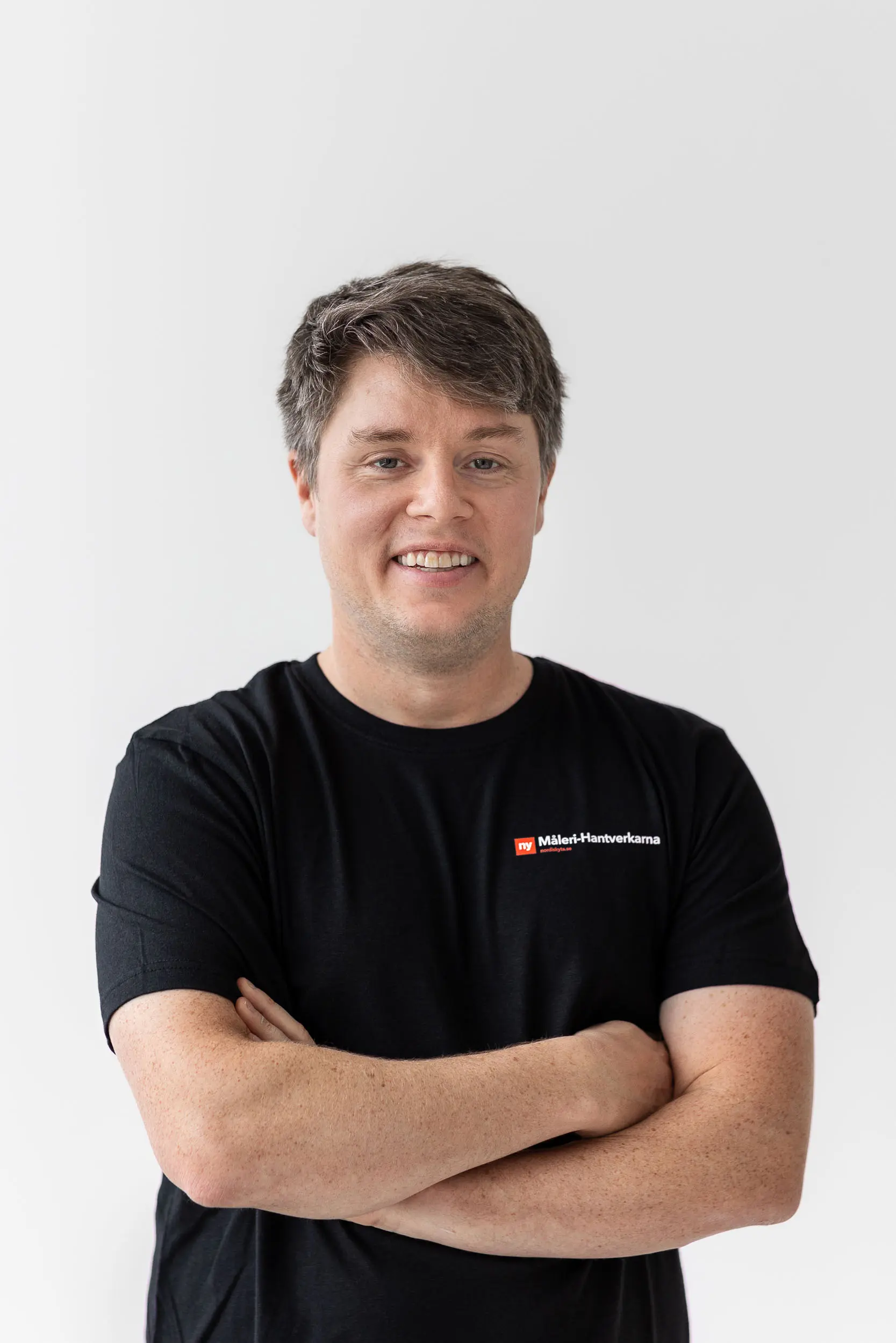 En person med kort brunt hår ler med armarna i kors, klädd i en svart t-shirt med en liten logotyp med texten Makita Husqvarna på vänster bröst, står mot en vit bakgrund.