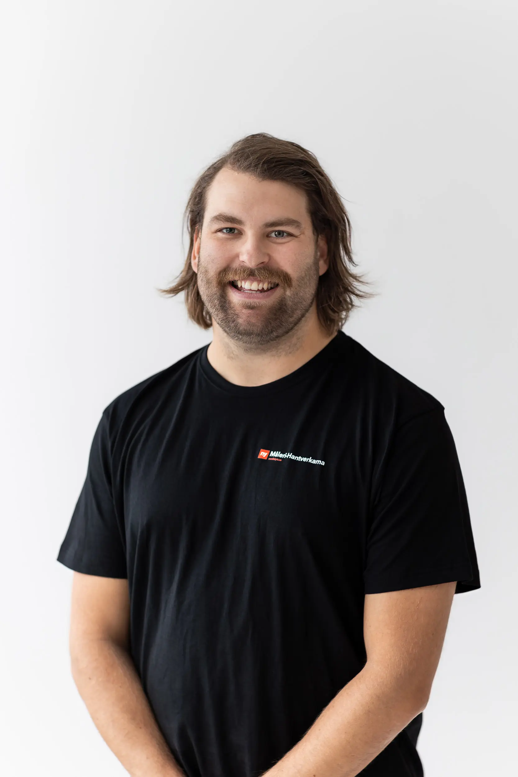 En man med axellångt brunt hår och skägg ler mot kameran. Han har på sig en svart t-shirt med en liten färgglad logotyp på vänster bröst och står mot en vit bakgrund.