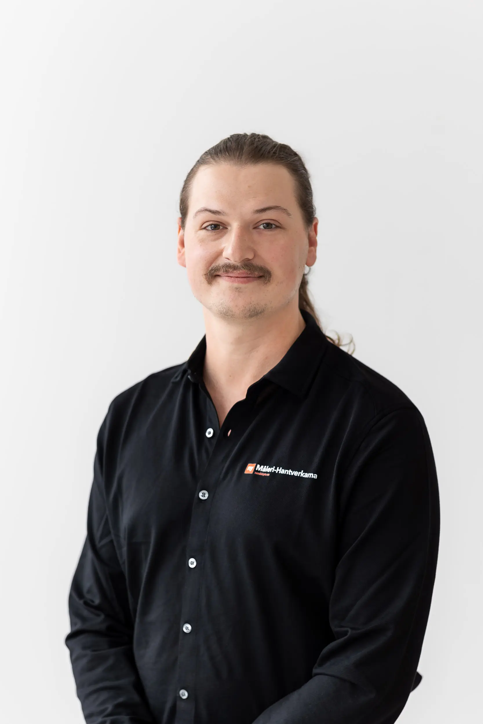 En person med långt, ljusbrunt hår som är bakåtkammat och mustasch står och ler, klädd i en svart skjorta med knappar och Maler-Handwerkskammarens logotyp, mot en vit bakgrund.