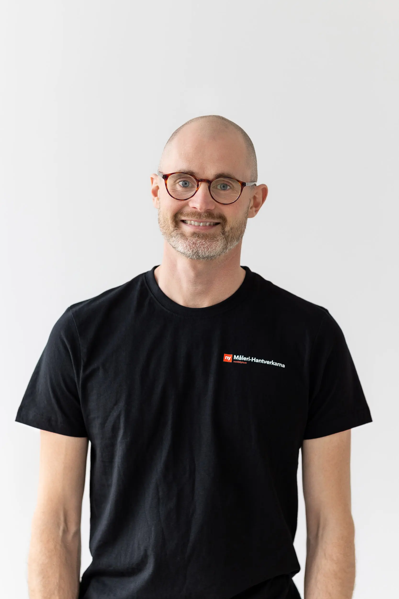 En leende man med glasögon, skägg och rakat huvud bär en svart t-shirt med en liten rödvit logotyp. Han står mot en vanlig ljus bakgrund.