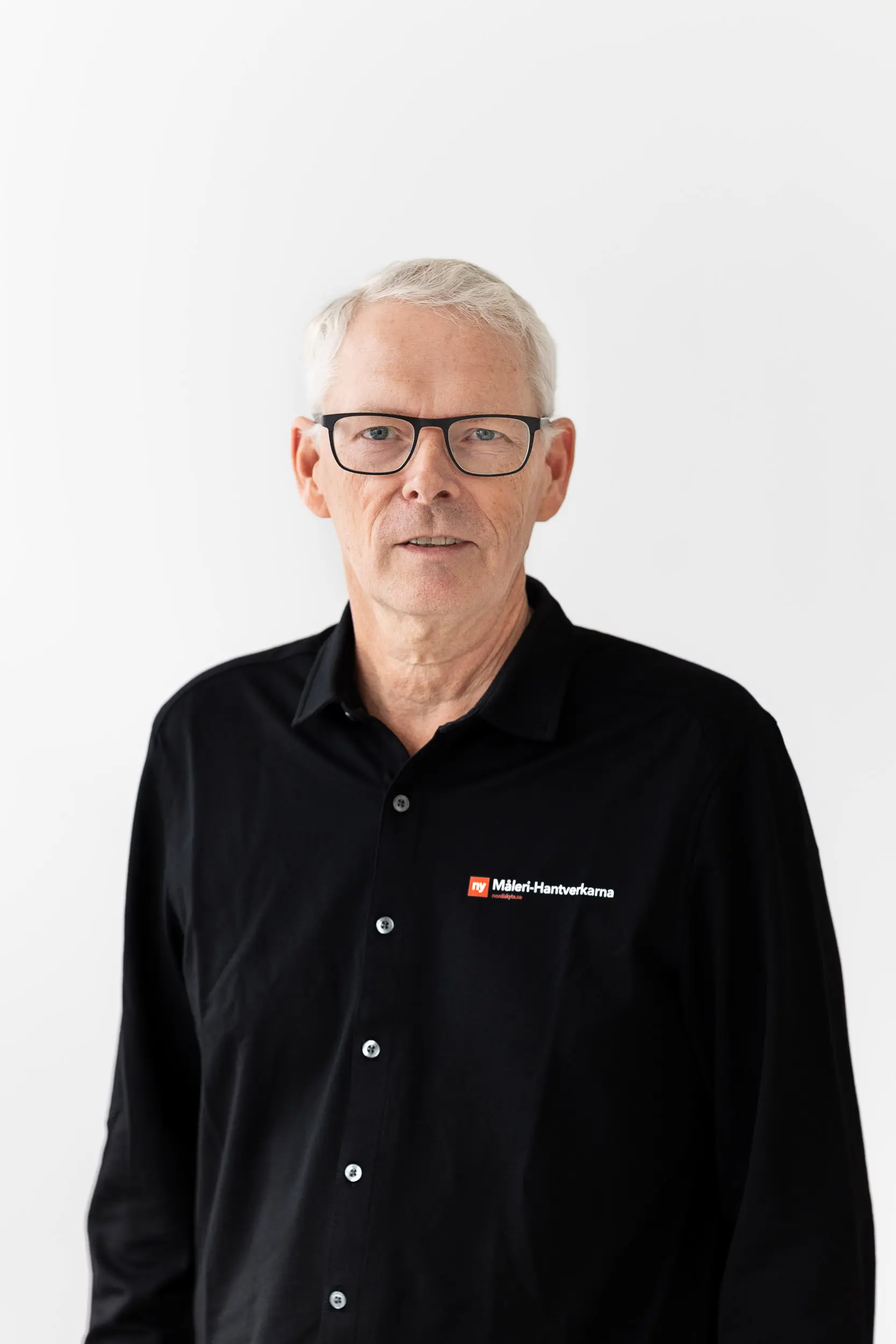 En man med kort grått hår och glasögon, klädd i en svart skjorta med knappar och Maler+Handwerkeramas logotyp, står framför en vit bakgrund.