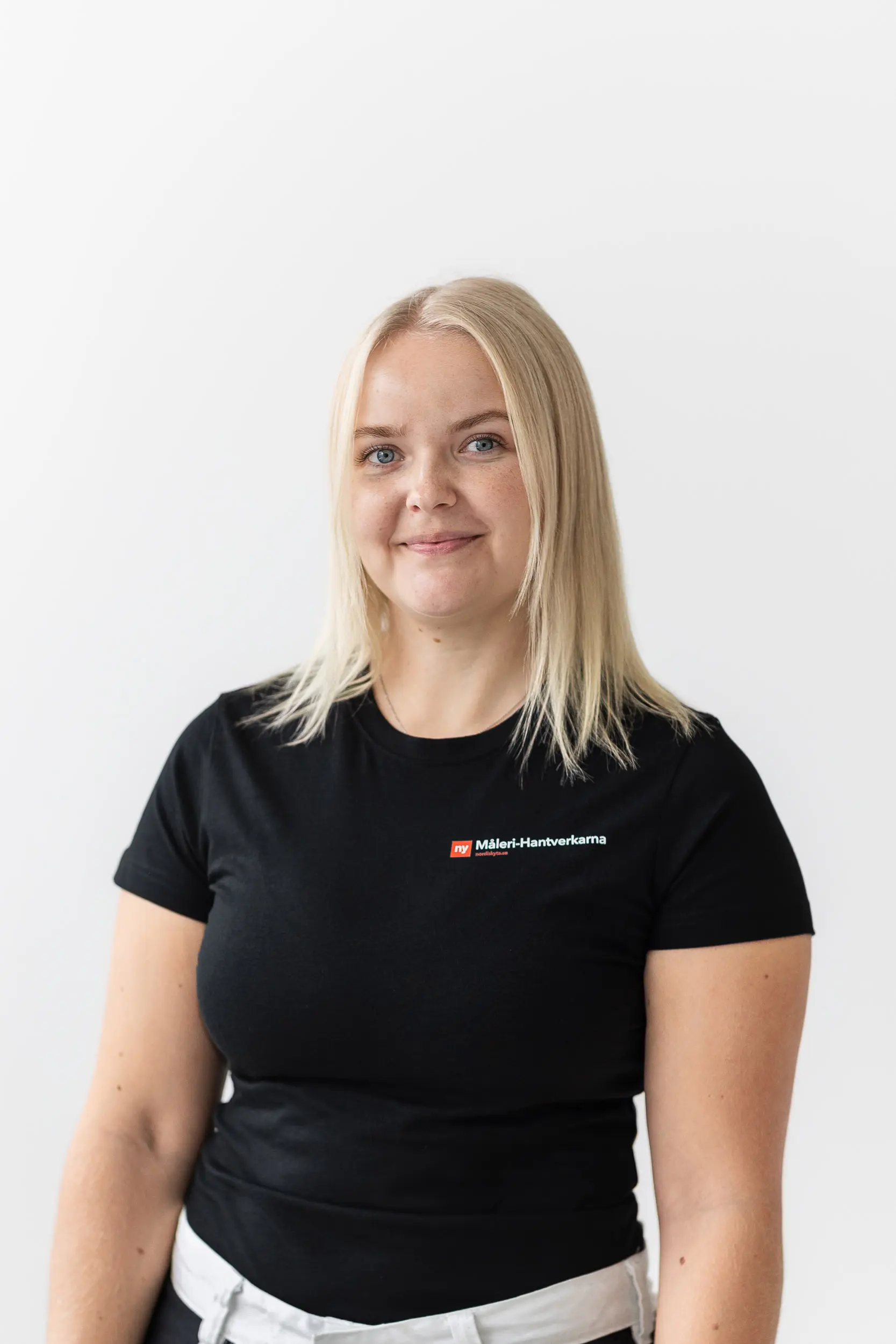 En kvinna med rakt blont hår, klädd i en svart t-shirt med en rödvit logotyp, står mot en vit bakgrund och ler mjukt mot kameran.