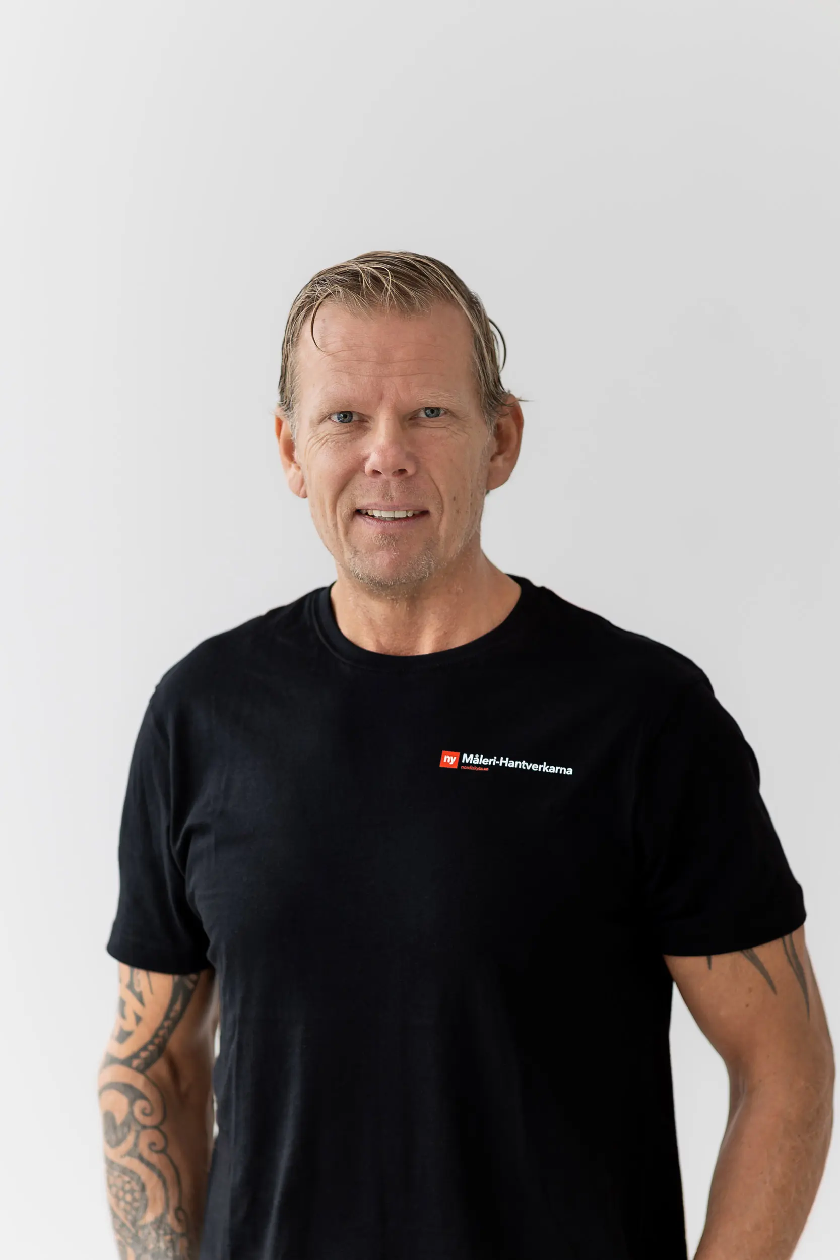 En man med kort, ljust hår och en tatuerad arm har på sig en svart T-shirt med en rödvit Maler-Hantverkvarna-logotyp och står mot en vit bakgrund.