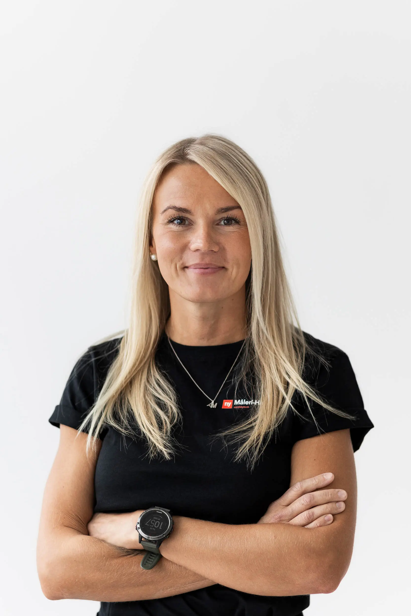 En kvinna med långt blont hår står mot en vit bakgrund, klädd i en svart t-shirt med en logotyp och en svart klocka, och ler med armarna i kors.