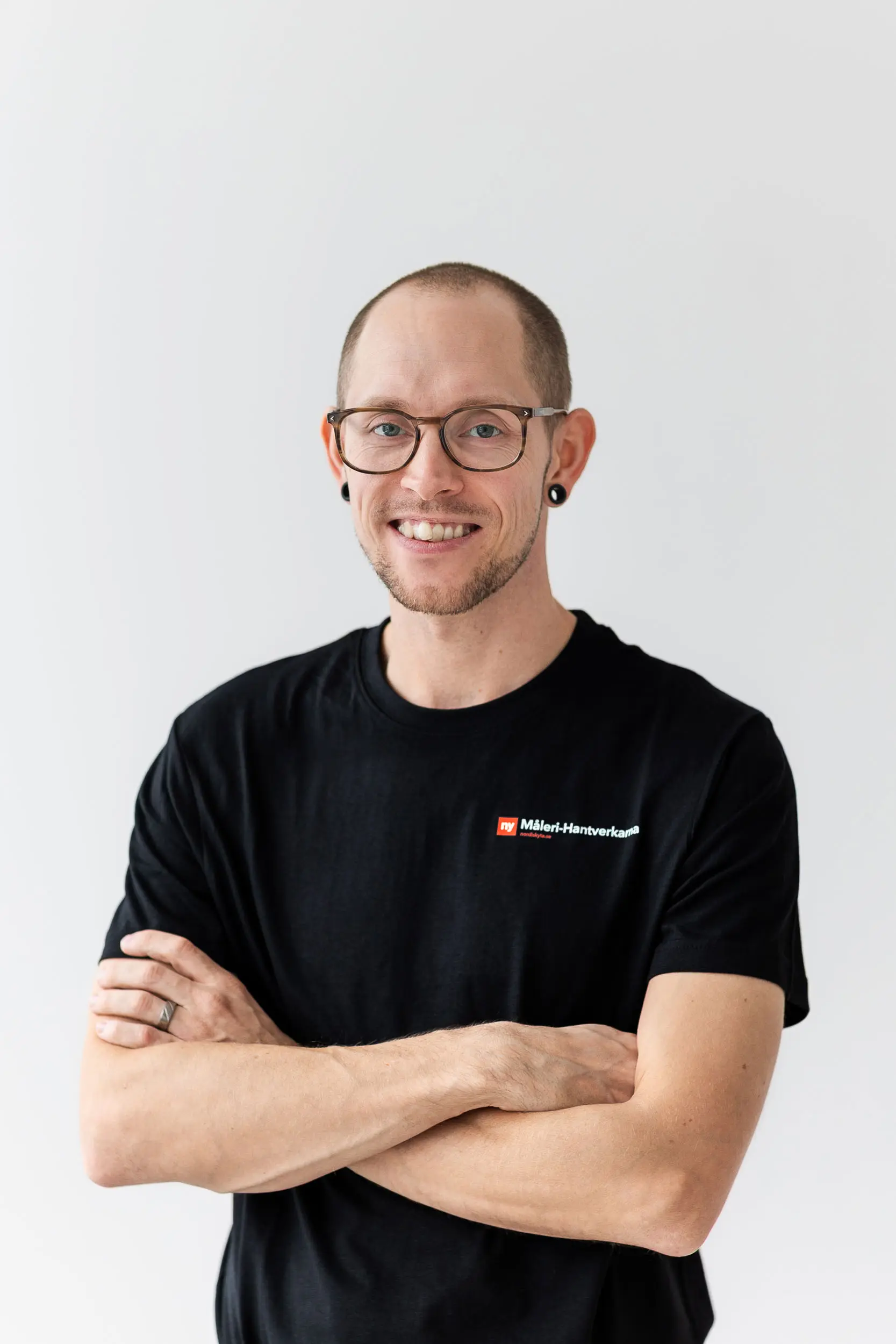 En leende person med glasögon och örhängen står med armarna i kors, iklädd en svart t-shirt med en liten rödvit logotyp på en ljus bakgrund.