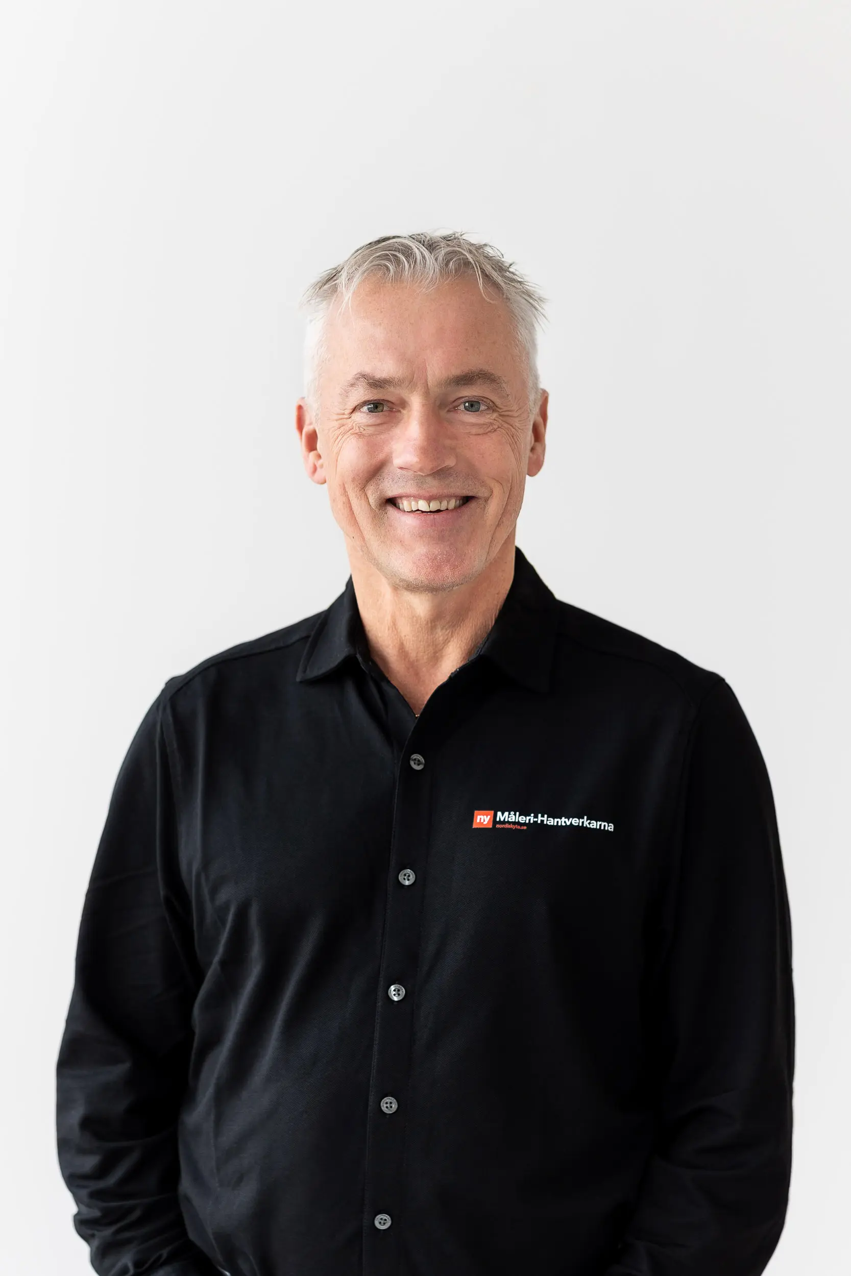 En leende äldre man med kort grått hår, klädd i svart skjorta med Mölnlycke Health Care-logotypen, står mot en vit bakgrund.