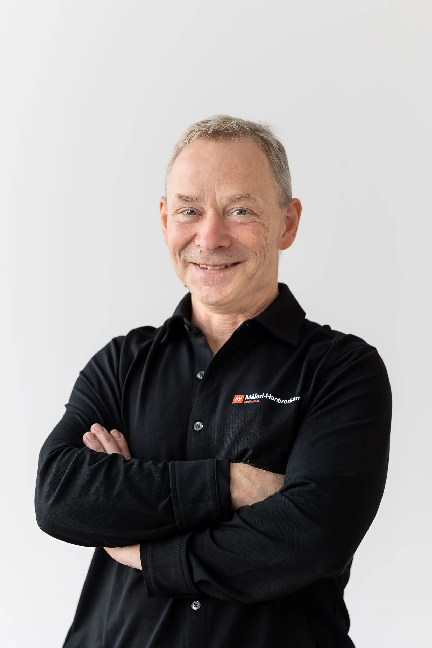 En medelålders man med kort grått hår ler stående med armarna i kors, iklädd en svart långärmad skjorta med en röd och vit företagslogotyp på bröstet, mot en vit bakgrund.
