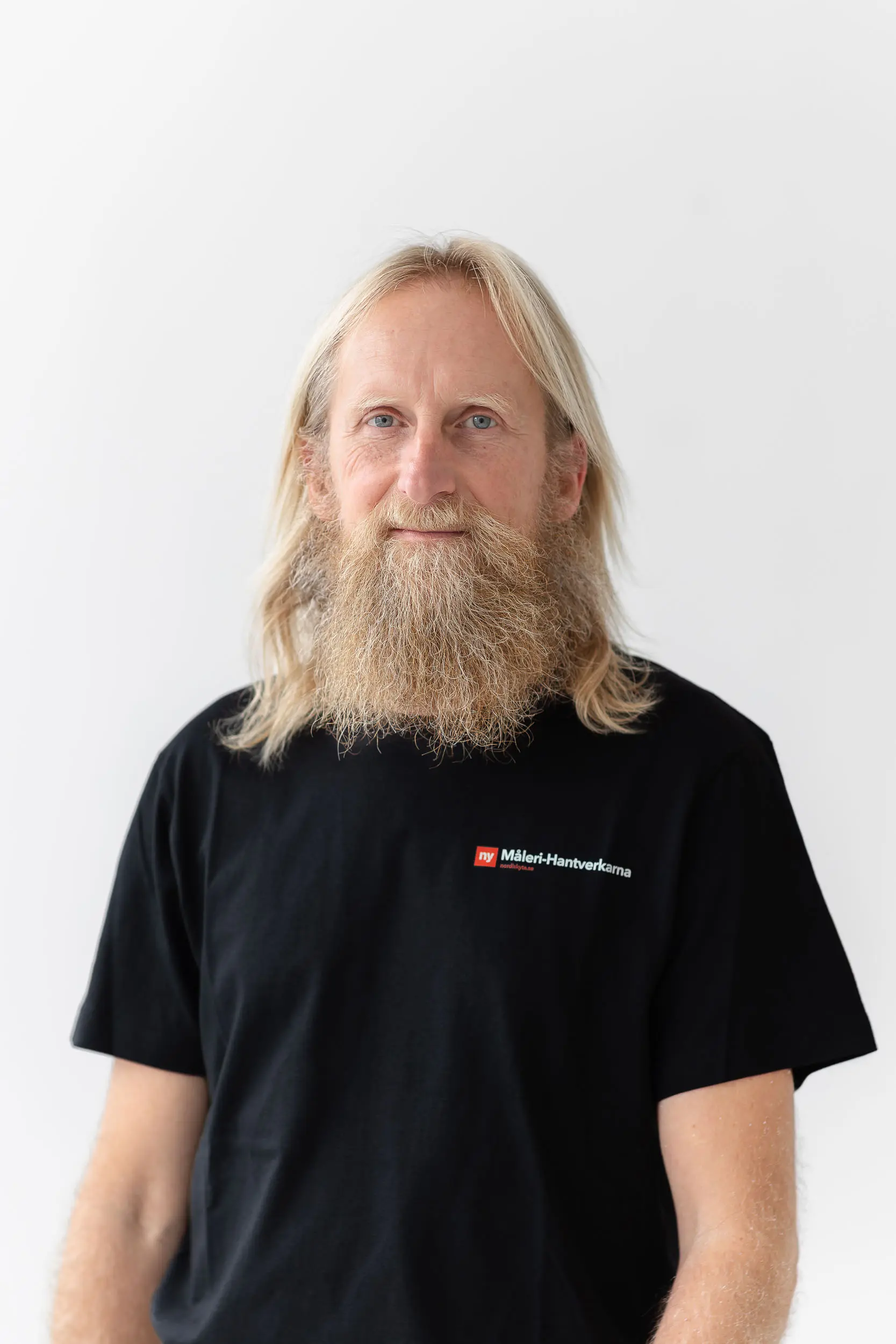 En man med långt blont hår och tjockt skägg står mot en vit bakgrund och har på sig en svart t-shirt med en liten rödvit logotyp på bröstet.