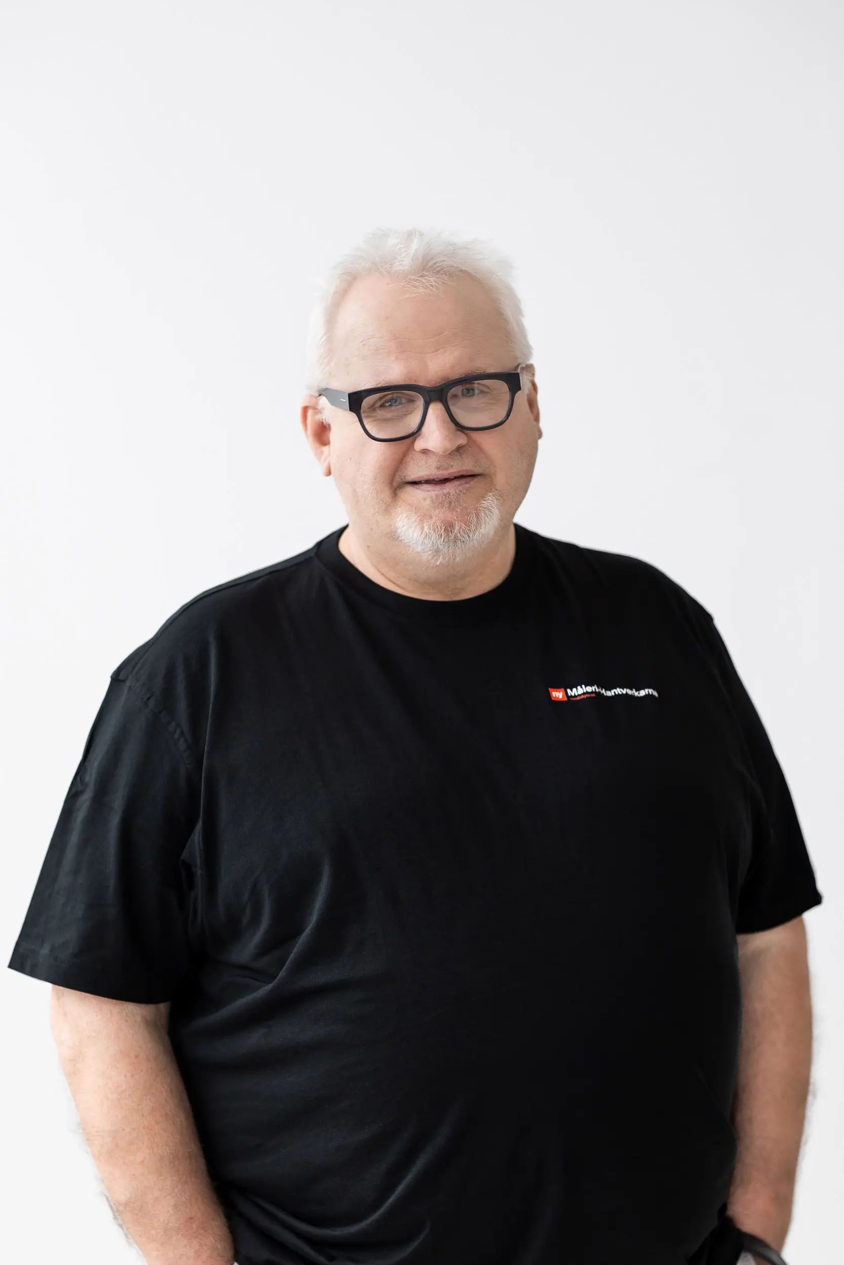 En man med kort vitt hår, glasögon och getskägg bär en svart T-shirt med en liten logotyp på vänster sida. Han står mot en vit bakgrund och ler lite mot kameran.