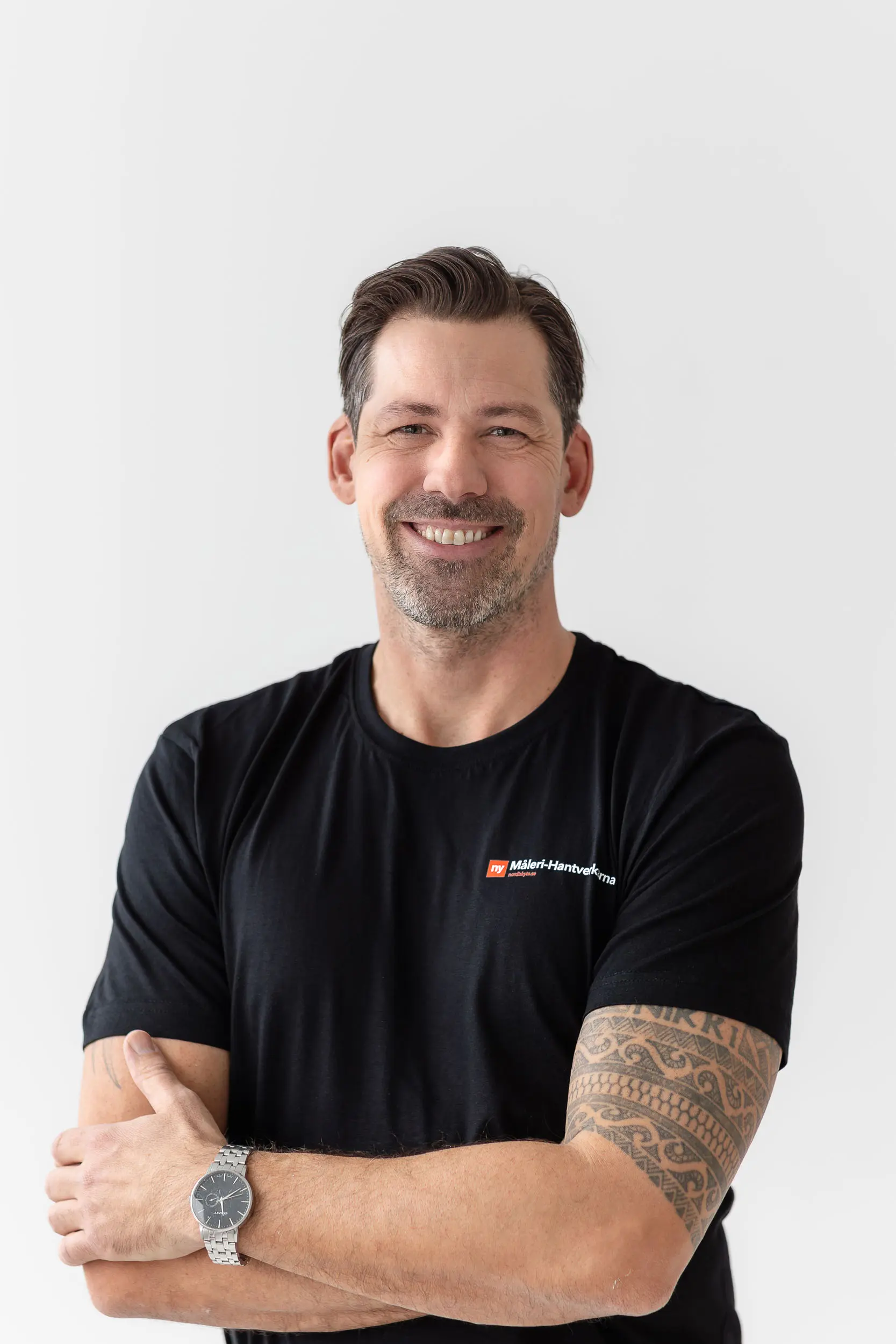 En leende man med kort brunt hår och skägg står mot en vit bakgrund. Han bär en svart T-shirt med en liten logotyp, har en tatueringsärm på vänster arm och en klocka i silver. Hans armar är korsade.