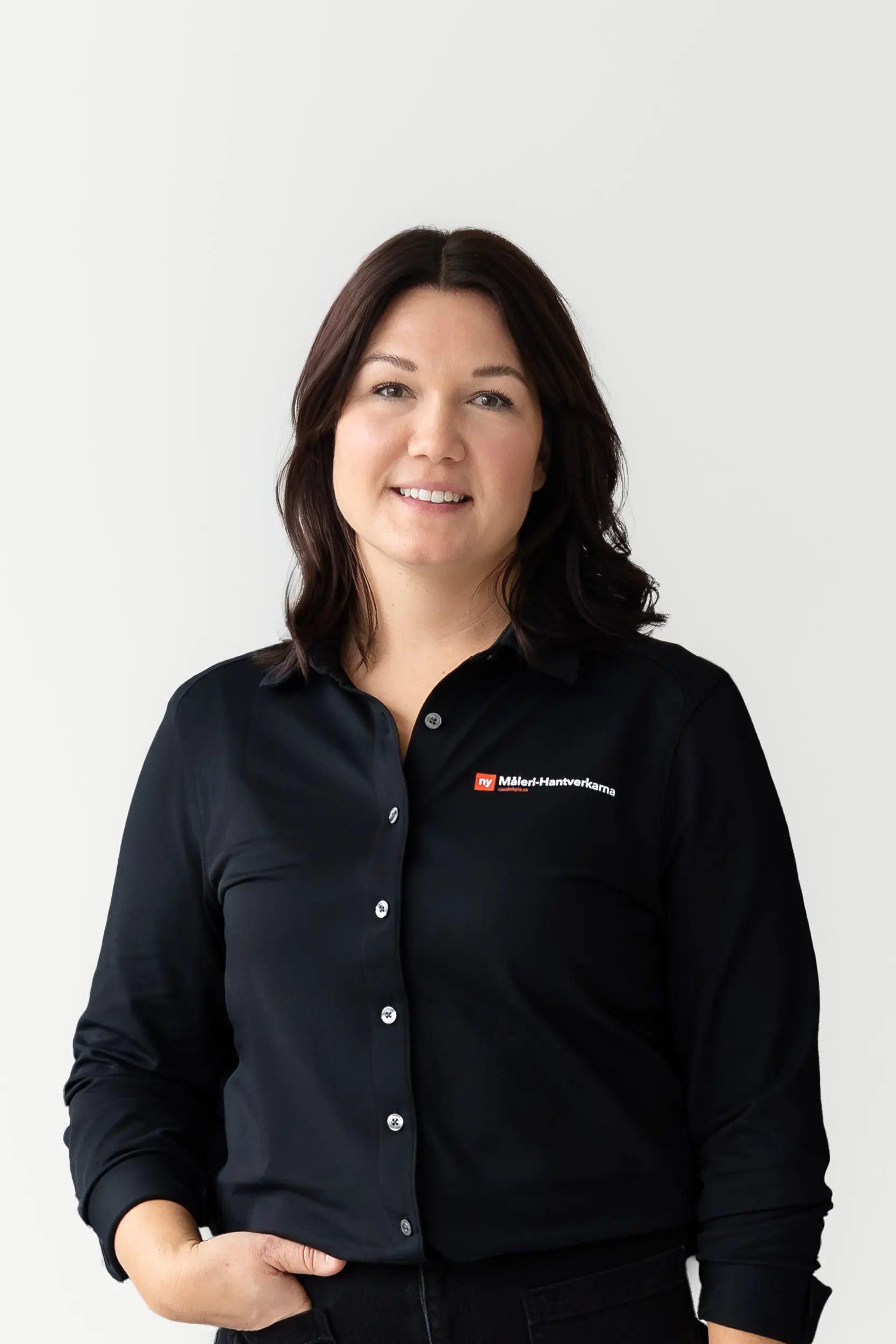 En kvinna med medellångt mörkt hår står och ler mot en ljus bakgrund. Hon bär en svart skjorta med knappar och en liten rödvit logotyp på vänster sida där det står Bilder & Hemleveranser.