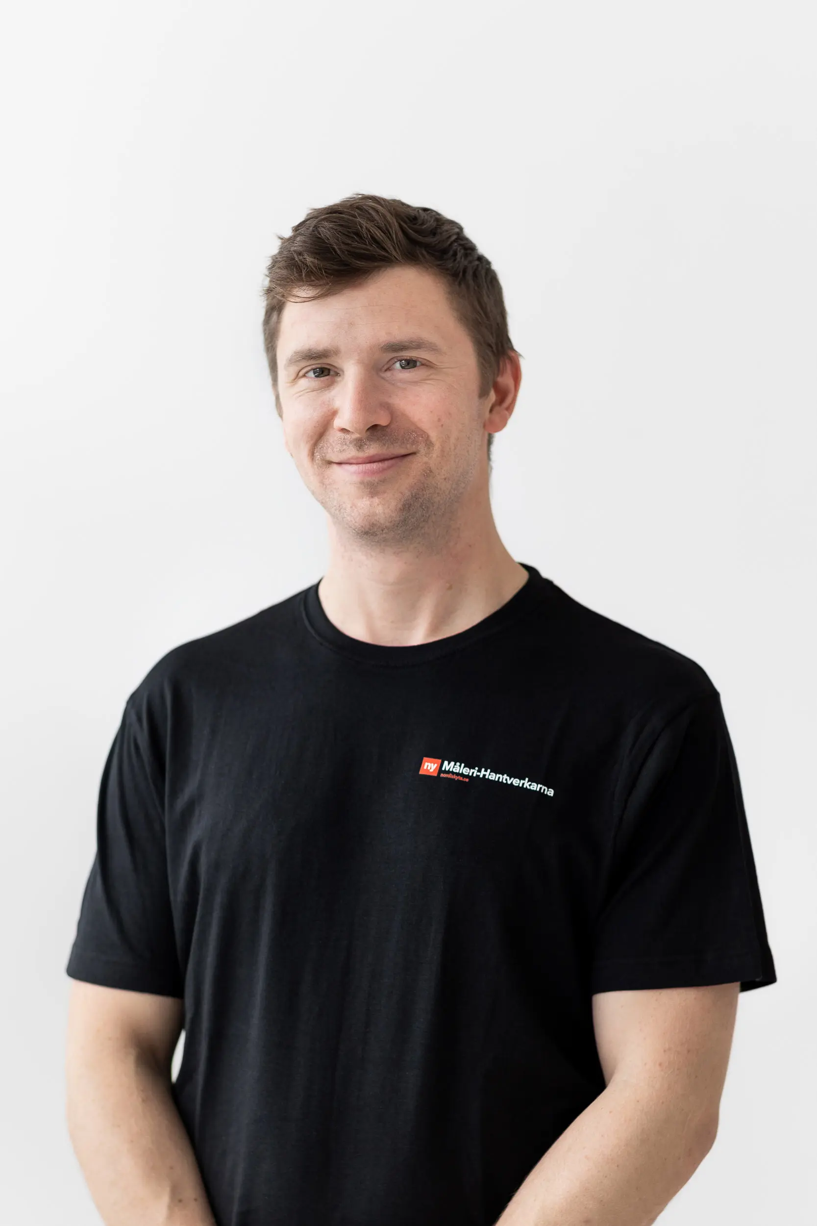 En man med kort brunt hår, klädd i en svart t-shirt med en rödvit logotyp och text, står mot en vit bakgrund och ler mot kameran.