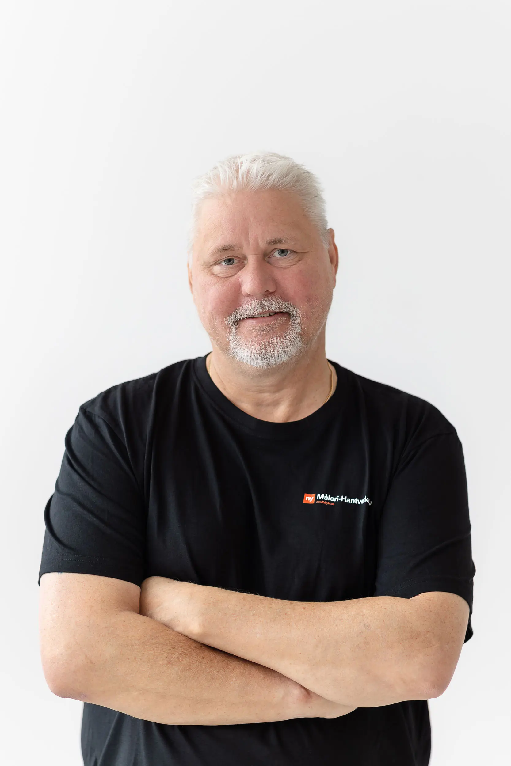 En äldre man med vitt hår och skägg står mot en vit bakgrund, klädd i en svart t-shirt med en liten rödvit logotyp på bröstet, med armarna i kors och ett vänligt leende mot kameran.
