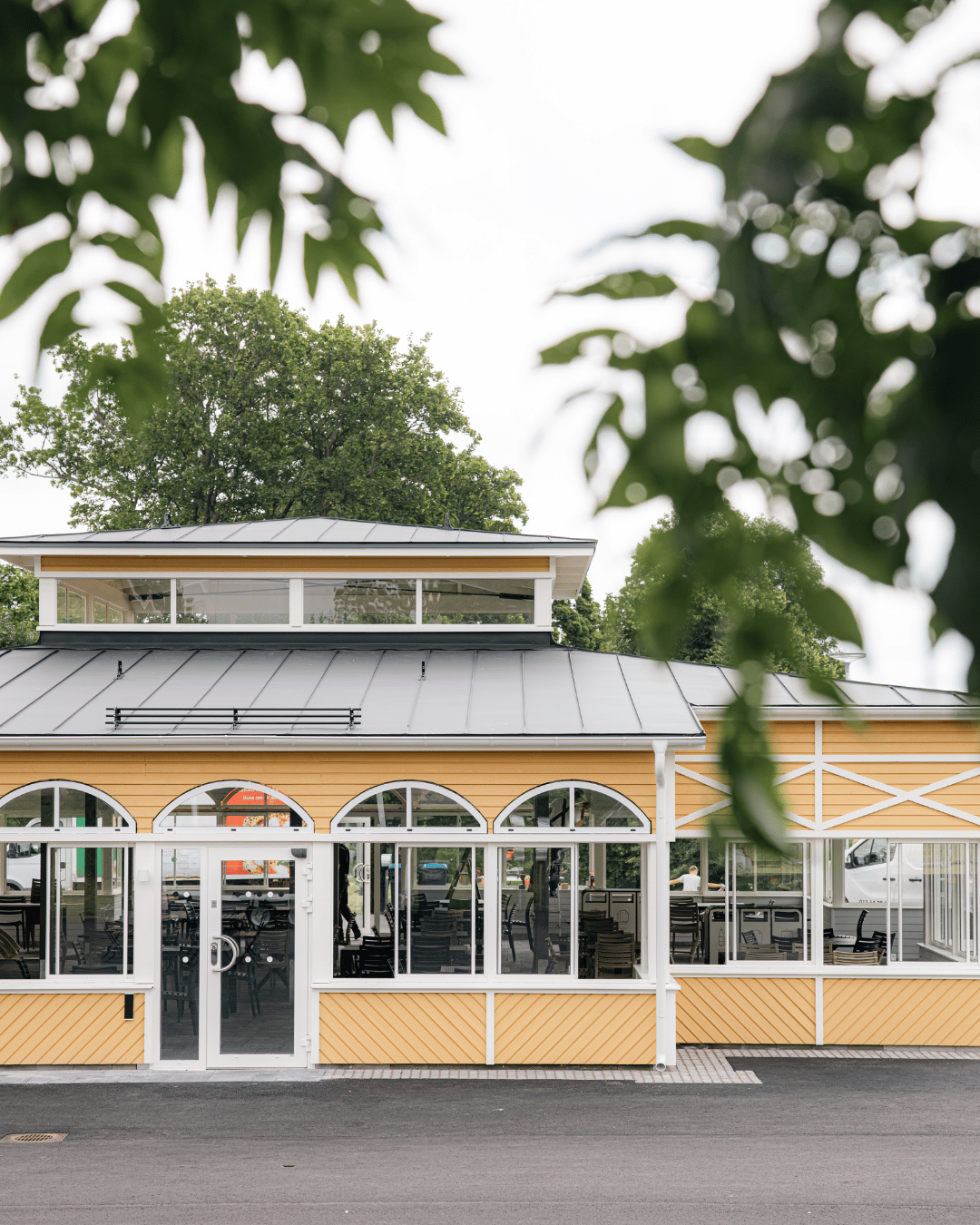 En gul byggnad med välvda fönster och ett plåttak omges av gröna trädgrenar. Genom fönstren syns bord och stolar utomhus.