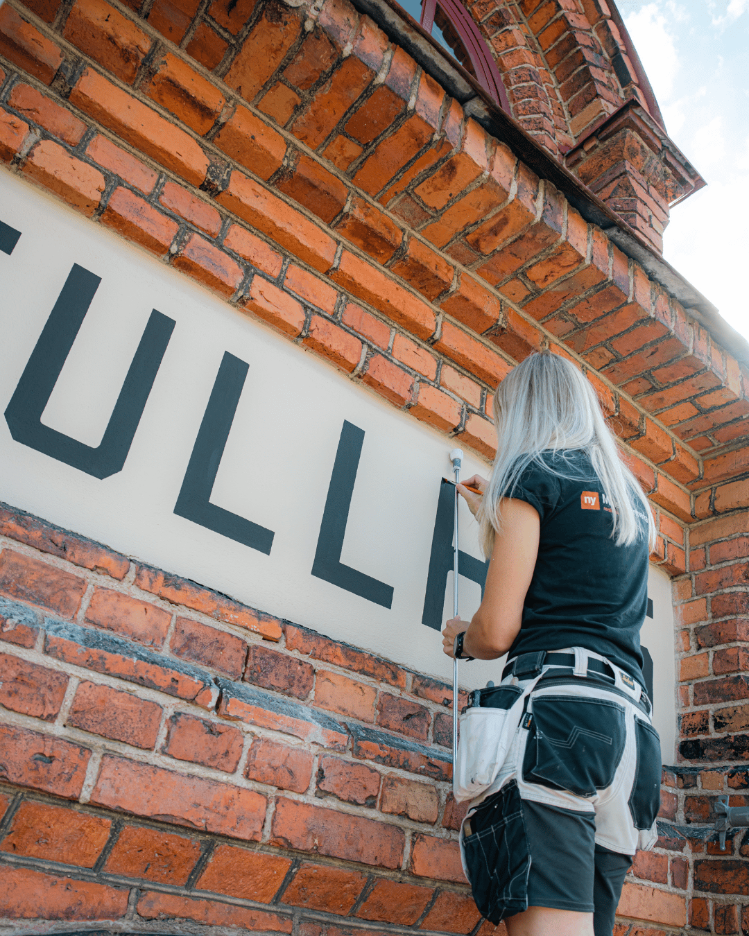 En person med långt blont hår målar stora svarta bokstäver på en vit skylt som sitter fast på en röd tegelbyggnad. Personen bär ett verktygsbälte och en svart skjorta.
