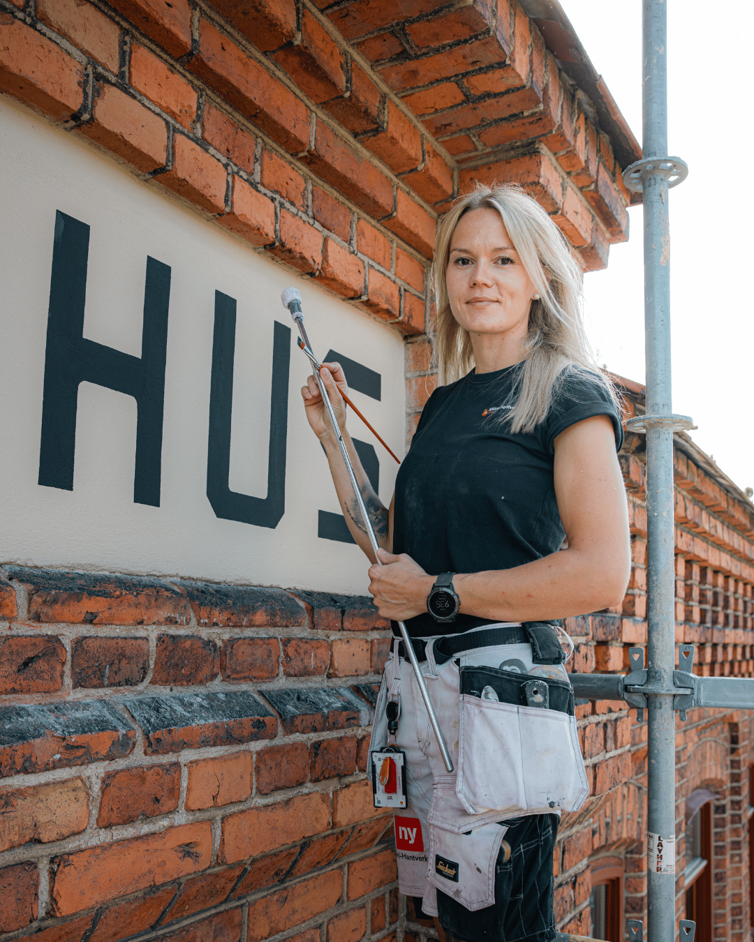 En kvinna står på en byggnadsställning intill en tegelbyggnad, med en målarrulle i handen, och målar stora svarta bokstäver på en krämfärgad skylt. Hon har på sig en svart tröja och ett verktygsbälte och tittar in i kameran.