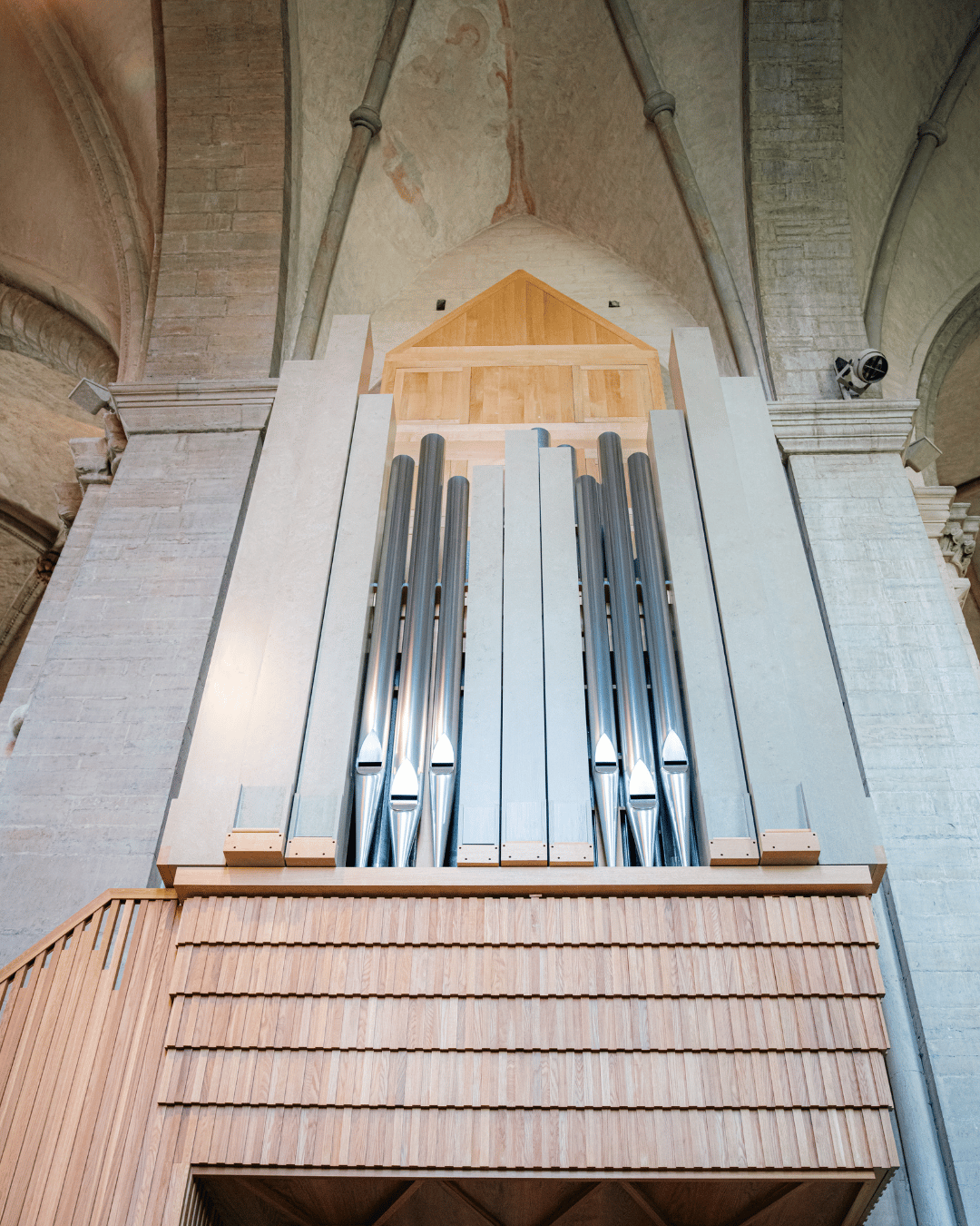 En stor piporgel med silverpipor och en träbas är inbyggd i en stenvägg i kyrkan under ett välvt tak.
