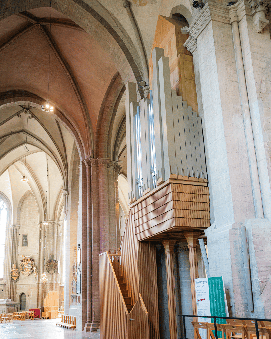 En stor piporgel med höga silverfärgade pipor och detaljer i trä står inne i en katedral med höga välvda tak och stenpelare. En trätrappa leder upp till orgelkonsolen.