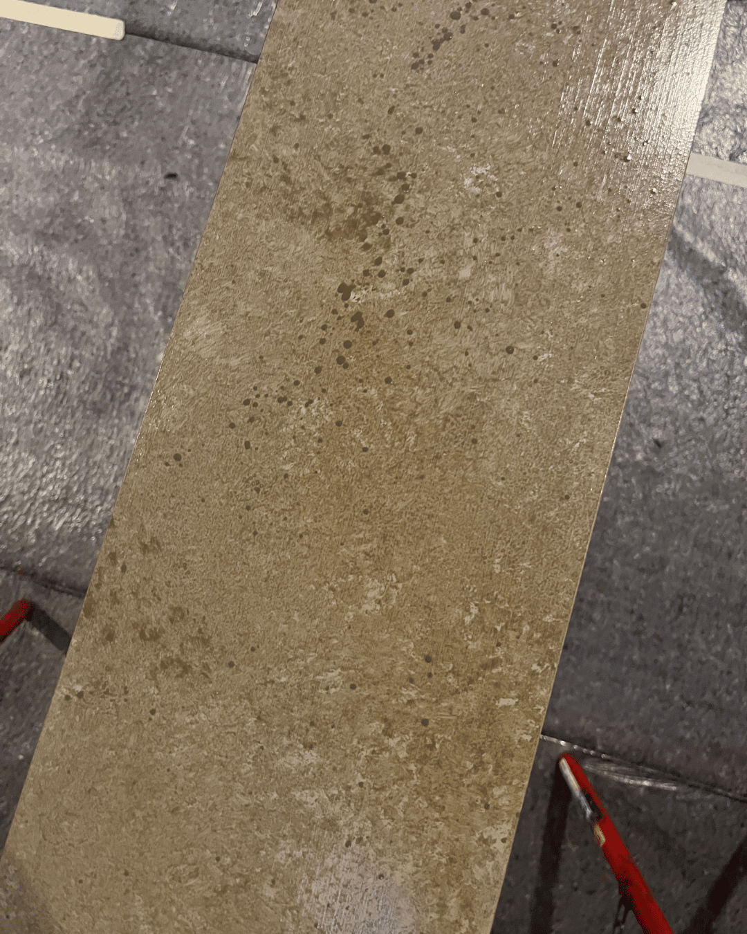 En närbild av en rektangulär kakelplatta med en strukturerad, brunaktig yta, där ojämna, fläckiga mönster syns. Kakelplattan vilar på röda stöd, med ett grått golv och vita linjer synliga i bakgrunden.