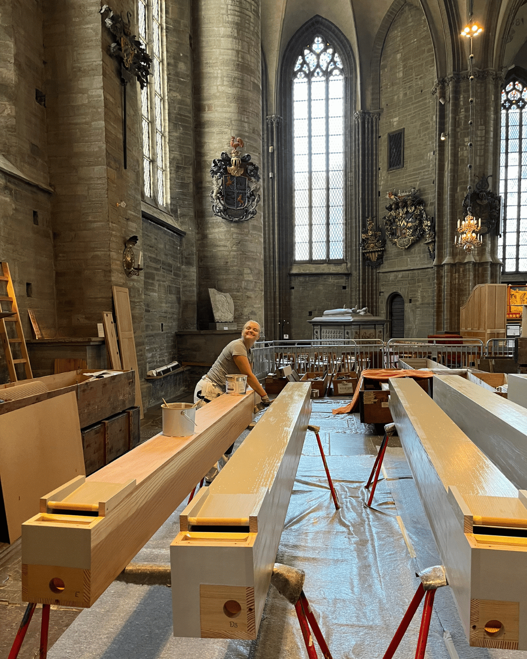 En person målar långa träbjälkar inne i en stor, historisk kyrka med höga välvda fönster, utsmyckade dekorationer och stenväggar.