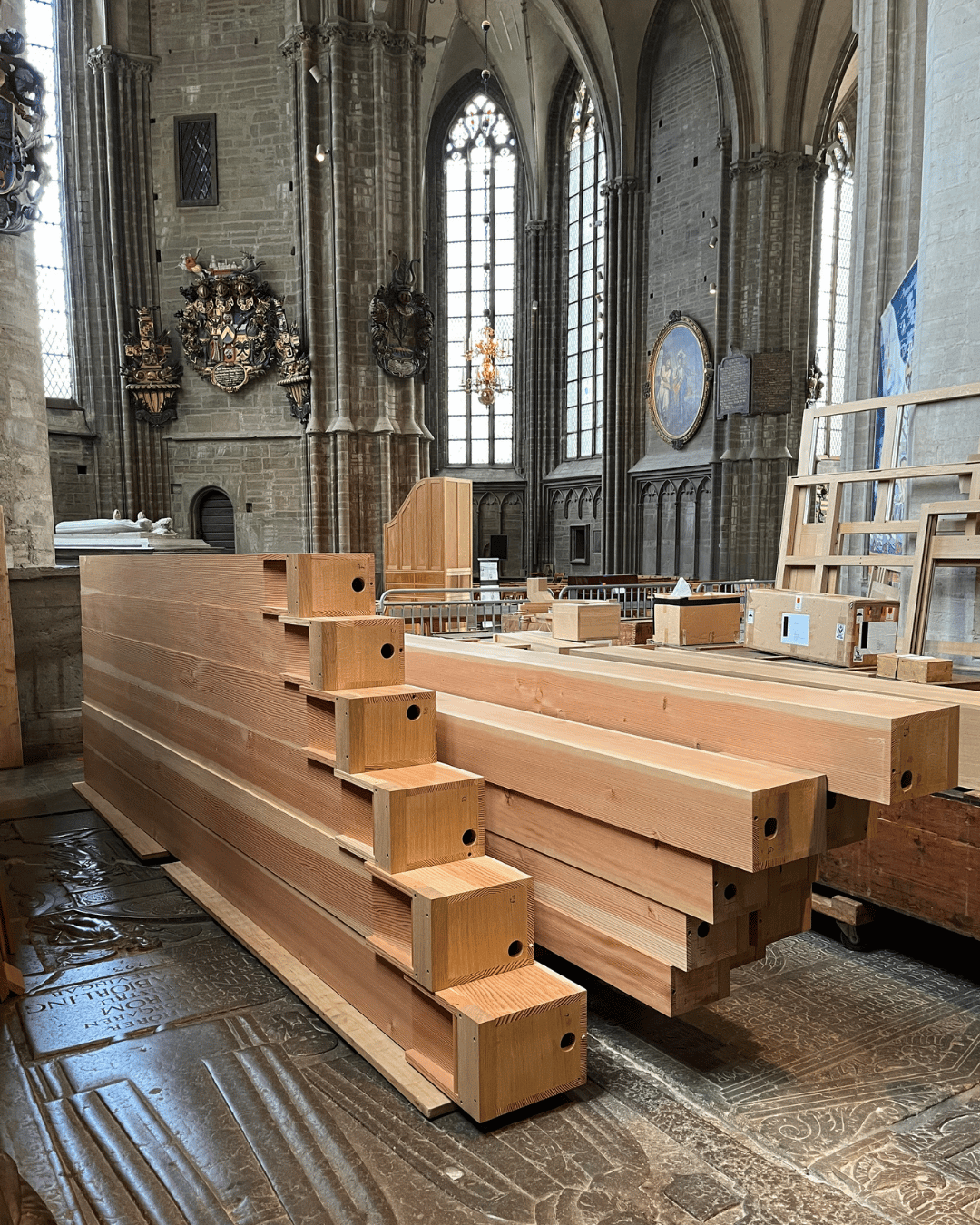Stora träbjälkar med fyrkantiga ändar är staplade inuti en historisk stenkyrka med höga välvda fönster, vapendekorationer och intrikat stenarbete. Byggmaterial syns i bakgrunden.