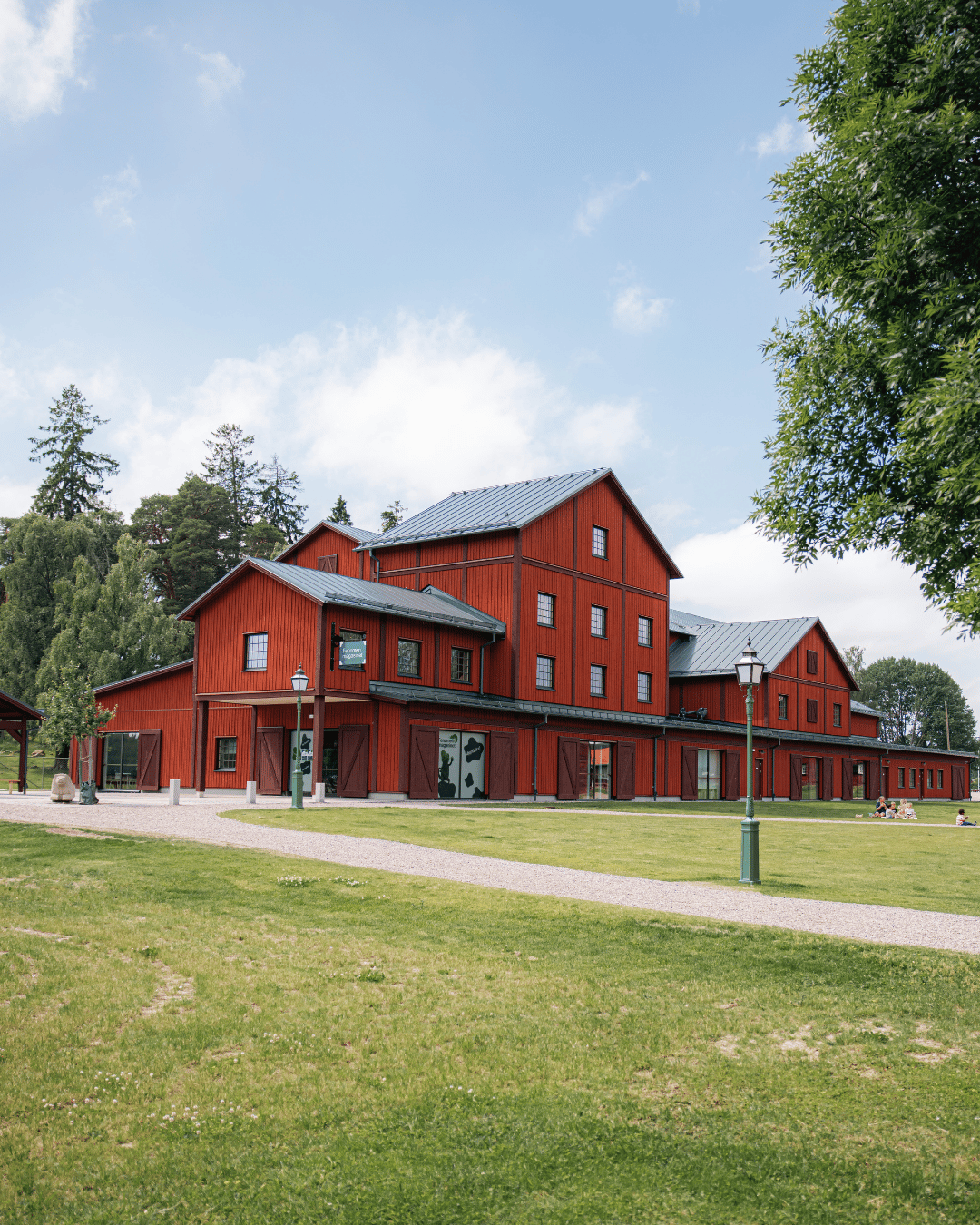 En stor röd träbyggnad med gröna detaljer står på en gräsbevuxen gräsmatta, omgiven av träd och en delvis molnig himmel.