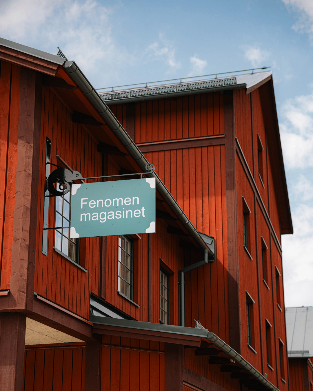 En blågrön skylt med texten ”Fenomen magasinet” hänger på en röd träbyggnad med sadeltak, med en delvis molnig himmel i bakgrunden.