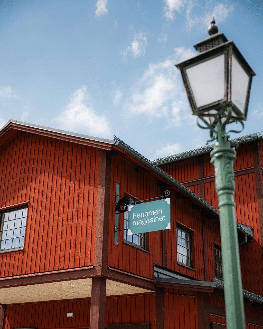 En grön gatlykta står framför en röd träbyggnad med en skylt där det står ”Fenomen magasinet”, mot en blå himmel med spridda moln.