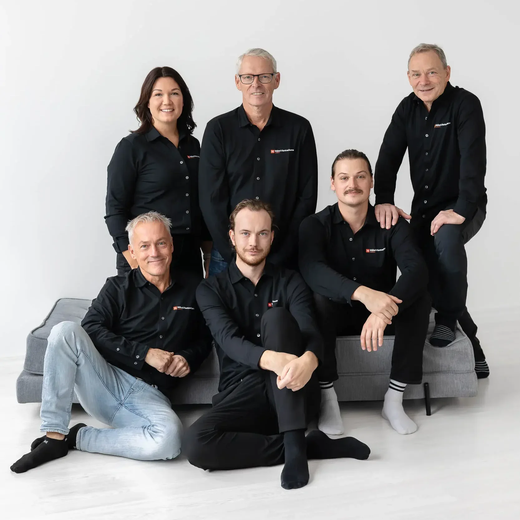 Sex vuxna personer i matchande svarta skjortor poserar tillsammans i ett ljust, minimalistiskt rum. Tre sitter i en grå soffa och tre står bakom dem, alla ler mot kameran mot en vit bakgrund.